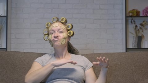 Funny woman with facial mask from cucumbers and in curlers dancing watching music channel on TV while sitting on couch.