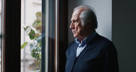 An elderly thoughtful smiling man is looking out a window of his house in the morning.