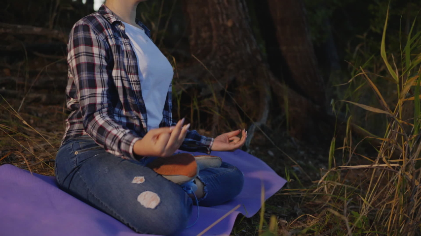 Teenage girl sitting on yoga mat and meditating in forest. Beautiful girl sitting in lotus position on mat and meditating with closed eyes in forest in the evening