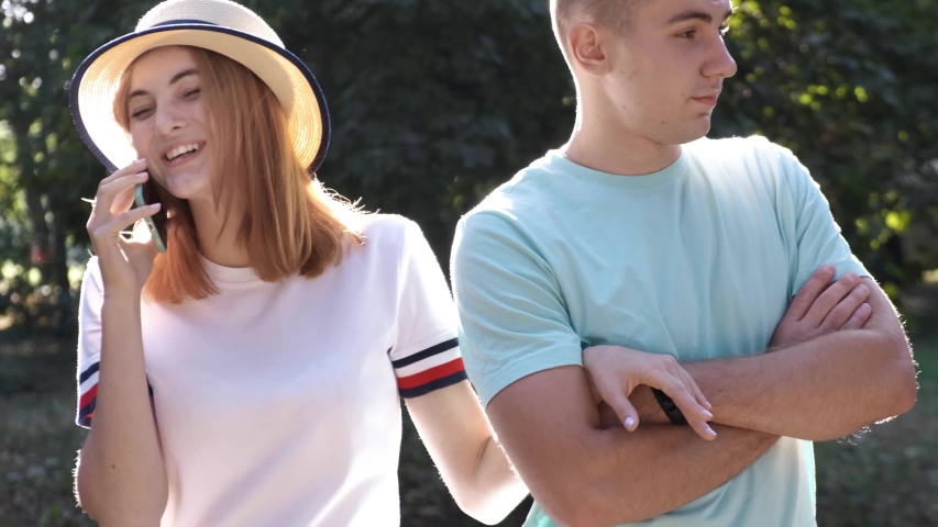 Portrait of pretty positive teenage girl with red hair wearing straw hat talking happily on mobile phone while her boyfrient is waiting bored beside.
