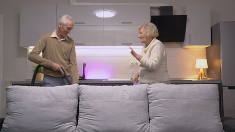 Cheerful old couple dancing together with vacuum cleaner and rag, happiness