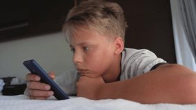 Closeup view of serious attentive face of cute caucasian kid using modern mobile telephone laying in bed at home. Real time 4k video footage.