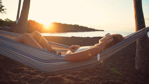 Woman contemplating sunset by the beach from her hammock. Young female relaxing in a hammock by the beach 