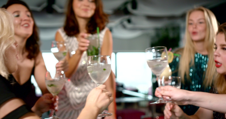 Two fateful girls in bright shiny dresses hang out and have fun at a party in a bar-club. They drink from the same glass with an alcoholic cocktail from different tubes. Royalty-Free Stock Footage #1043707486