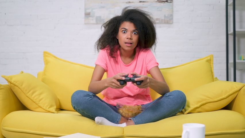 Young afro american woman sitting on the sofa and playing video game. Victory emotion.