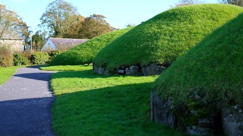 handheld wide shot of the archeological enclosure of Knowth, Boyne Valley Tombs with green grass covered tombs and an ancient stone cottage in the background on a sunny blue sky day and no people.