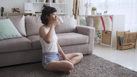 Beautiful young asian japanese woman drinking tea while resting at home. lovely smiling female in sleepwear sitting on carpet in morning living room enjoy cup of coffee. bright modern house interior.