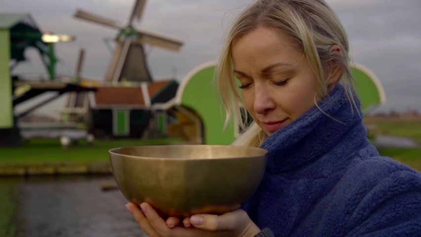 Portrait of a fair-haired girl in a blue coat, she is on the background of the river and three mills. He holds a metal bowl in his hands.
