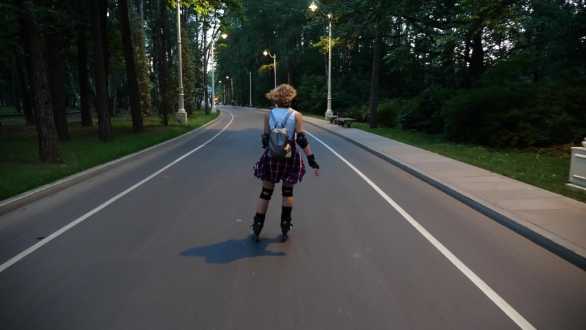 A young curly blonde girl is quickly roller skating on a track in the park in the evening. The camera follows an active athletic woman on roller skates and in protective gear.