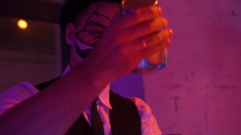  RUSSIA, TULA - NOVEMBER 1, 2019 Halloween young man in white shirt black yolk and hat beautifully and quickly shakes shaker with an alcoholic drink with a make-up of an orange mask and a white skull