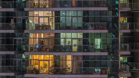 Rows of glowing windows and balconies with people in the interior of apartment building at night. Modern skyscraper with glass surface. Concept for business and modern life. Zoom in