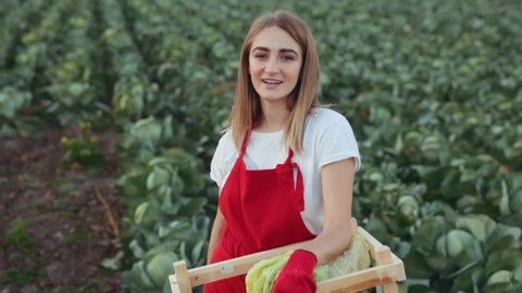 Portrait of cheerful european young woman farmer in red apron holding wood box with cabbage, looking into the camera and smiling, feeling good, happy. Close up, slow motion. Shoot on ARRI ALEXA.