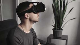 Young bearded man wearing headset of virtual reality in office sitting on a sofa. Exploring VR world and all what he saw in headset.