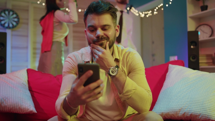 Unsociable bearded guy using mobile phone checking online news using application getting bored at night party. Young people dancing having fun on background. | Shutterstock HD Video #1043826376
