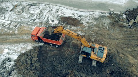 Aerial view from above of a construction site with excavators and trucks working. A loader puts stones and sand into a truck.