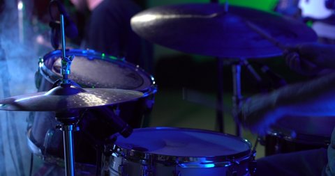 A drummer plays on a dark stage in the fog and light of lanterns, in soundcheck time. The average plan is a hand with a stick, percussion, cymbals, a snare drum.