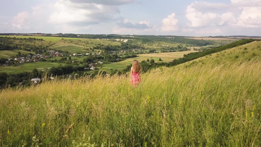 Yong woman with long hair in red dress walking in summer field with tall green grass on rural hill environment.