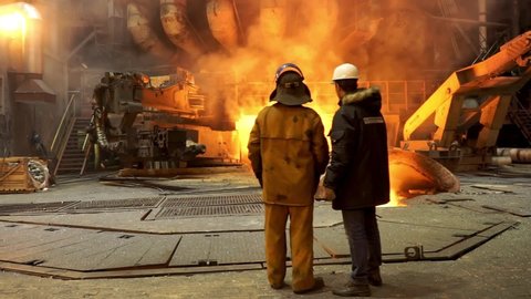 Steelworkers standing near a blast furnace with sparks at the metallurgical plant, heavy industry concept. Stock footage. View inside of hot shop of the steelmaking factory.