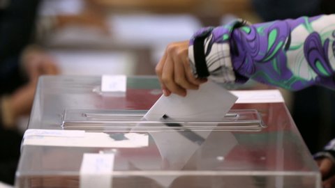 A woman casts her envelope vote into a transparent plastic ballot box. Human hand voting.