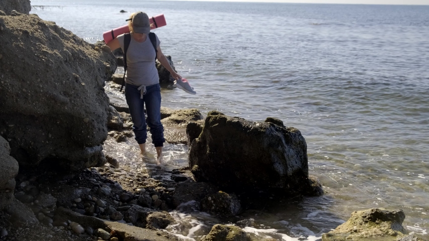 girl traveler passes through water barefoot walking along rocky shore near sea. woman tourist walks along edge of rocky shore washed by sea.