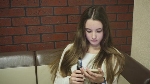 Vaping teenager with problem skin. Young cute caucasian girl smoking an electronic cigarette and surfing in a smartphone in the vape bar. Deadly bad habit. Vaping activity. Close up. Social problem.