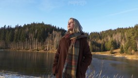 blonde in a coat and scarf stands by the lake in the forest