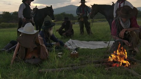 Cowboys wearing on cowboy hat and cowboy boots stopped camping and rested while boiling the coffee flask above the fire flame and some people are rested and sleep at the horse saddles. Arkivvideo
