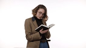 Smiling beautiful brunette young woman in glasses and brown jacket isolated over white background in studio. People sincere emotions, lifestyle concept. Reading book, excited and thoughtfully smiling