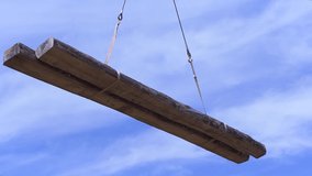 Two wooden beams being transported by the crane on blue cloudy sky background. Clip. Bottom view of logs in the air.