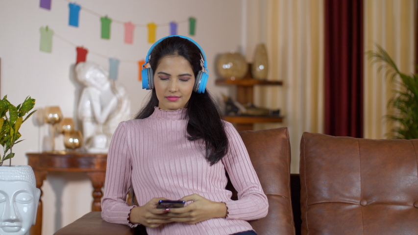 Closeup shot of an Indian girl listening to her favorite songs using her blue headphones. Young beautiful female enjoying music with her Bluetooth headphones while spending leisure time at home - w...