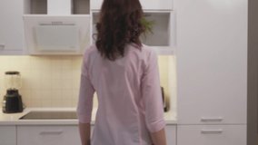 Charming slender woman in pink blouse and jeans opening refrigerator full of organic food and taking pitcher with orange juice. Young girl with brown hair drinking fresh beverage at home.