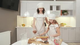 Charming young lady in kitchen apron and hat teaching cute female child to make homemade ginger cookies. Mother and her daughter flattening dough before baking in modern kitchen