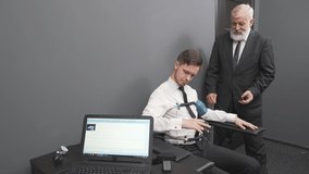 Mature man with grey beard in black suit attaching sensors on fingers and body to young guy that sitting in black chair. Handsome man passing check with polygraph detector in grey room.