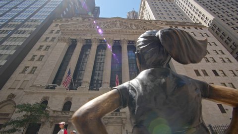 NEW YORK CITY, USA – JUNE 26, 2019: Fearless Girl bronze sculpture facing The New York Stock Exchange building on Wall Street NYC, panning motion view, sunny summer day