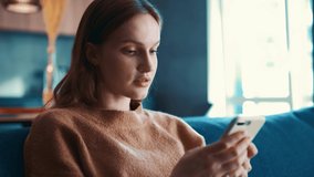 Beautiful caucasian woman in beige sweater sitting on a blue sofa and using smartphone or cell phone in modern room. Tapping, scrolling, watching video, content, bloggs.