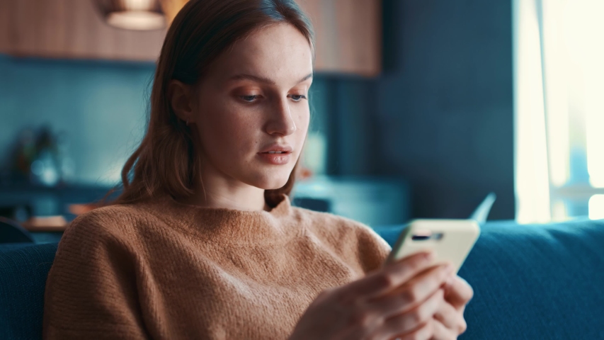 Beautiful caucasian woman in beige sweater sitting on a blue sofa and using smartphone or cell phone in modern room. Tapping, scrolling, watching video, content, bloggs. | Shutterstock HD Video #1043934169