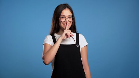 Smiling asian woman with long hair holding finger on her lips over blue background. Gesture of shhh, secret, silence. Close up.