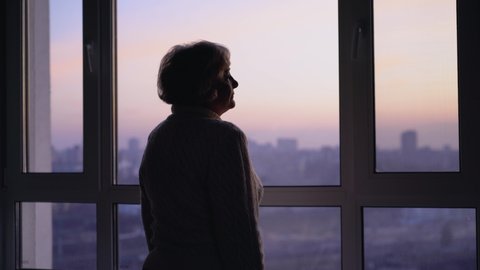 Lonely senior woman sadly looking in window, missing family at retirement home