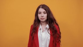 Beautiful brunette young woman in fashion white shirt and red jacket keeping hands on jacket, staying sideways isolated on orange background in studio. People sincere emotions, lifestyle concept.