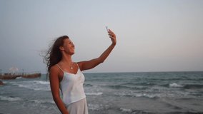 Happy woman using smartphone to video call on beach, waving hello and giving thumbs up
