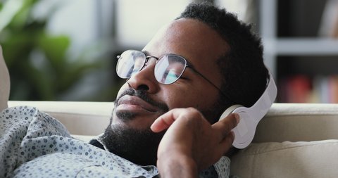 Serene calm young african man wear wireless headphone relax with eyes closed meditate lying on sofa listen lounge music audio book course feel peace of mind no stress on couch at home, close up view