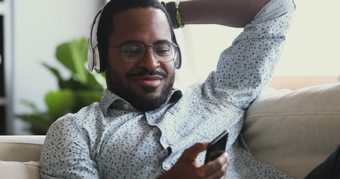 Happy carefree mixed race guy wearing wireless headphones listening to music on smartphone sit on sofa, relaxed smiling young african man enjoying mobile player audio sound lounge on couch at home