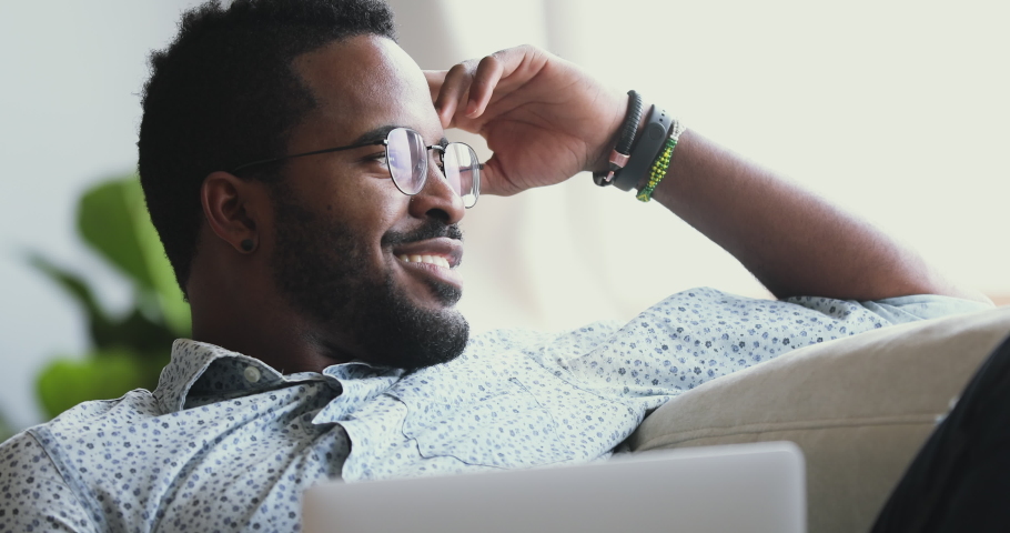 Happy afro american man wear glasses relax with laptop looking away dreaming resting on sofa at home, smiling young mixed race guy lounge on couch with notebook feel satisfied concept, close up view Royalty-Free Stock Footage #1043980060