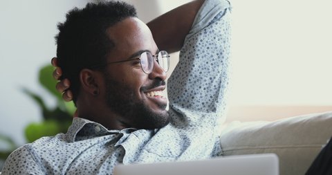 Happy afro american man wear glasses relax with laptop looking away dreaming resting on sofa at home, smiling young mixed race guy lounge on couch with notebook feel satisfied concept, close up view
