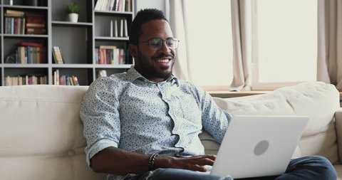 Happy young african male student using laptop notebook surfing social media relaxing at home, smiling mixed race man enjoying easy e learning browsing internet on modern technology device sit on sofa