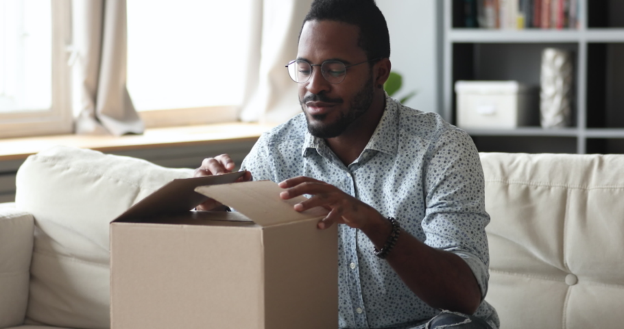 Happy african man customer open cardboard box receive gift online purchase in postal parcel shipping delivery service concept, satisfied mixed race male consumer unpacking package sit on sofa at home | Shutterstock HD Video #1043980084