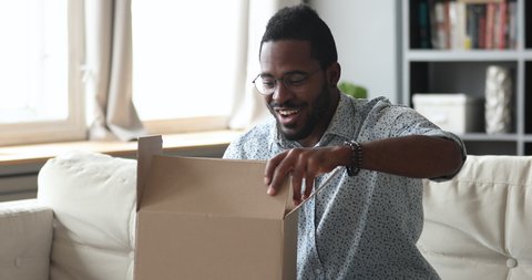 Happy african man customer open cardboard box receive gift online purchase in postal parcel shipping delivery service concept, satisfied mixed race male consumer unpacking package sit on sofa at home