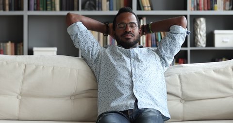 Happy serene healthy young african mixed race man relax on comfortable sofa holding hands behind head enjoy no stress peace of mind concept feel calm breath fresh air dream lounge on couch at home