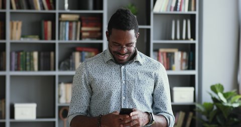 Smiling young hipster african businessman entrepreneur hold smart phone stand in office, happy mixed race millennial male student professional user using mobile app texting messages on cellphone tech