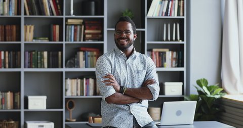 Confident african businessman entrepreneur teacher stand in modern office with bookshelves look at camera, smiling young mixed race professional posing in work space arms crossed, business portrait